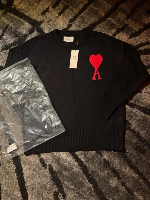 Svart stickad tröja från Ami Paris - Svart stickad tröja från Ami Paris med röd hjärtlogga och bokstaven A på bröstet. Tröjan har rund halsringning och ribbade muddar vid ärmslut och nederkant. Perfekt för en stilren och trendig look.