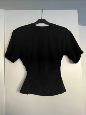 Svart figurnära topp med kort ärm - Snygg svart topp med korta ärmar och figurnära passform. Toppen har en markerad midja som ger en smickrande siluett och är tillverkad i ett mjukt material. Perfekt att styla med jeans eller kjol för en trendig look.