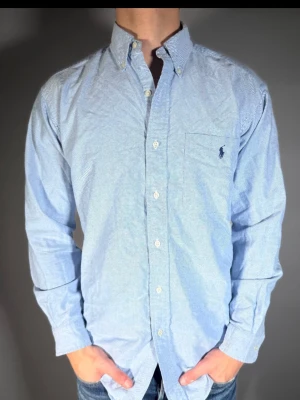 Ljusblå skjorta från Ralph Lauren - Klassisk ljusblå skjorta från Ralph Lauren med button-down krage och bröstficka med broderad logga. Skjortan har långa ärmar, vita knappar och är tillverkad i mjuk bomull. Perfekt för en clean och stilren look.