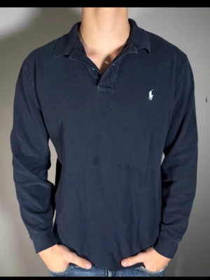 Mörkblå långärmad henley från Polo Ralph Lauren - Klassisk mörkblå långärmad henley från Polo Ralph Lauren med vit broderad logga på bröstet. Skjortan har krage och knappar vid halsen, tillverkad i mjuk bomull för en bekväm känsla. Perfekt till jeans eller chinos.