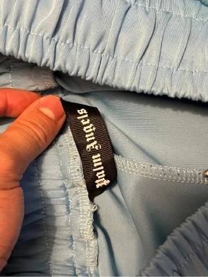 Ljusblåa mjukisbyxor från Palm Angels kommer med påse - Säljer ett par ljusblåa mjukisbyxor från Palm Angels med broderad logga på benet. Byxorna har resår i midjan och är gjorda i ett mjukt, glansigt material som känns riktigt skönt. Perfekta för en chill och trendig look.