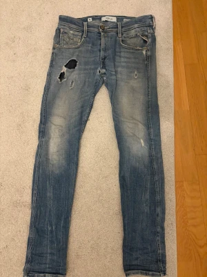 Replay anbass jeans - Säljer ett par blå jeans från replay med slitningar. Jeansen är slim fit och klassisk knappgylf. Materialet är denim i bomull och färgen är tvättad blå med ljusare partier. Nypris 1200 kr