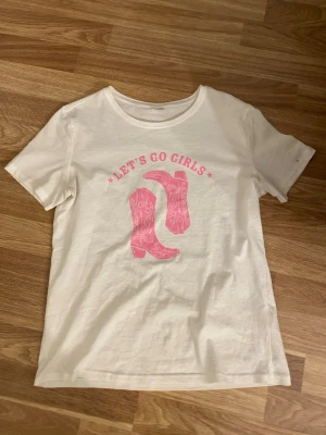 Vit t-shirt med rosa cowboyboots - Köpt på en butik i Kroatien! Vit t-shirt med tryck i rosa av cowboyboots och texten 'LET'S GO GIRLS' framtill. Klassisk rund hals och korta ärmar. Perfekt för dig som gillar statement-plagg med cool western-vibe.