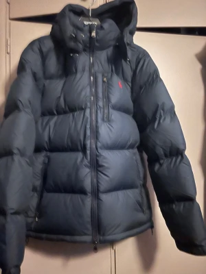 Marinblå dunjacka från Polo Ralph Lauren - Säljer en marinblå dunjacka från Polo Ralph Lauren med huva. Jackan har en röd logga på bröstet Perfekt för kalla vinterdagar och riktigt snygg! Helt ny med alla lappar kvar, inköpt på Väla på DO. Nypris 5999:-