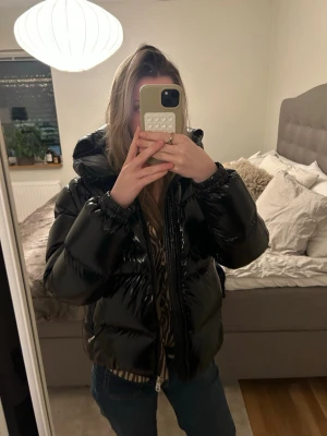 Glansig colmar pufferjacka - Säljer en svart, glansig colmar pufferjacka med huva och dragkedja framtill. Jackan har en oversized passform och elastiska muddar vid ärmsluten. Perfekt för kalla vinterdagar och ger en cool streetwear-look. (Finns galja att få med )