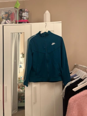 Blå ziptröja från Nike med ränder - Sportig blå ziptröja från Nike med vita ränder längs ärmarna och logga på bröstet. Tröjan har hög krage, dragkedja framtill och normal passform. Perfekt för en avslappnad och cool stil.