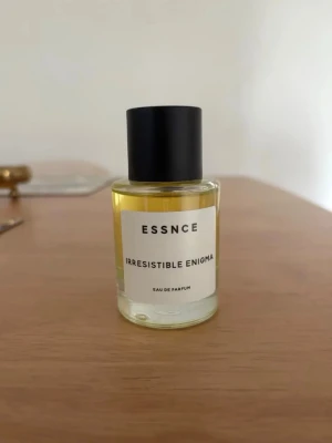 ESSNCE Irresistible Enigma parfym - En stilren parfymflaska från ESSNCE med doften Irresistible Enigma. Flaskan är cylindrisk i genomskinligt glas med gulaktig vätska och svart rund kork. Minimalistisk etikett med svart text. Perfekt för dig som gillar moderna och eleganta dofter.