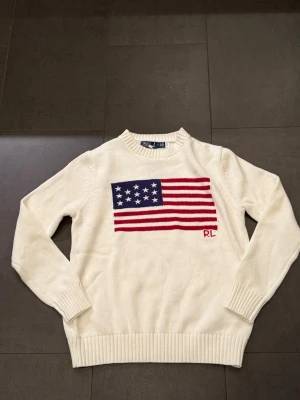 Vit stickad tröja från Ralph Lauren - Snygg vit stickad tröja från Ralph Lauren med amerikansk flagga i rött, vitt och blått på bröstet samt RL-broderi. Tröjan har rund halsringning, ribbade muddar och är tillverkad i mjukt material. Perfekt för dig som gillar klassisk och preppy stil.