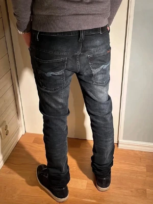 Svarta slim fit jeans från Nudie Jeans - Säljer ett par svarta slim fit jeans från Nudie Jeans med snygga slitningar och broderade detaljer på bakfickorna. Jeansen har klassisk femficksmodell och är tillverkade i stretchigt denimtyg för skön passform.