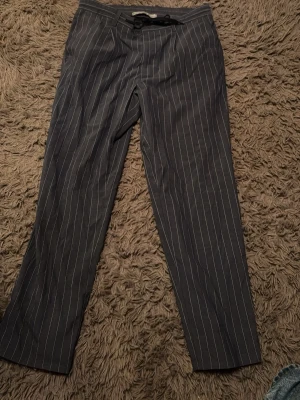 Randiga kostymbyxor från MNG MAN - Snygga mörkgrå kostymbyxor från MNG med vita smala kritstrecksränder. Byxorna har rak passform och dragsko i midjan för en avslappnad men ändå dressad look. Perfekta för dig som gillar stilrena detaljer och klassisk design.