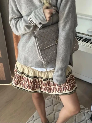 Beige kortkjol med volang och mönster - Supersöt beige kortkjol med volangkant och broderat mönster i rött och grönt. Kjolen har en avslappnad passform och elastisk midja för extra komfort. Perfekt att styla med en stickad tröja eller sneakers för en trendig look.