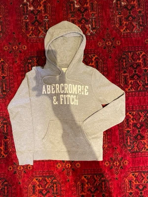 Abercrombie & Fitch Hoodie - Snygg grå hoodie från Abercrombie & Fitch med stor vit logga på bröstet. Klassisk känguruficka framtill och justerbar huva med dragsko. Mjuk bomullsblandning som känns skön mot huden. Perfekt för en avslappnad och trendig look. Undrar du något är det bara att skriva😁🙌