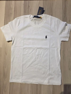 Vit t-shirt från Polo Ralph Lauren - Klassisk vit t-shirt från Polo Ralph Lauren med den ikoniska loggan broderad på bröstet. T-shirten har rund hals, korta ärmar och är gjord i mjuk bomull för en skön känsla. Perfekt basplagg till garderoben.