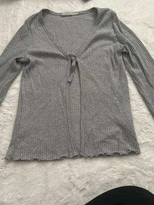 Grå ribbad omlottkofta med knyt - Säljer en grå ribbad omlottkofta med v-ringning och knyt framtill. Koftan har långa ärmar och är tillverkad i ett mjukt material som ger en skön passform. Perfekt att slänga på sig när du vill ha något enkelt men snyggt.