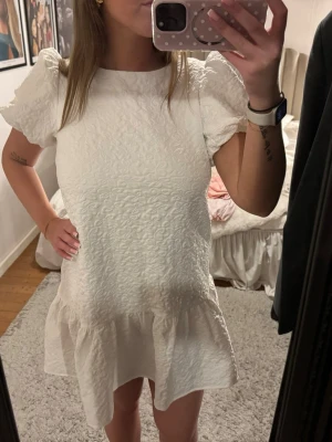 Studentklänning - Supersöt vit kort klänning med puffärmar och volang nertill. Klänningen har en strukturerad yta och rund halsringning. Perfekt till studenten