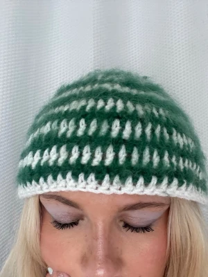 Grön och vit stickad mössa - Säljer en handvirkad mössa i grönt och vitt med breda ränder. Mössan är gjord i ett fluffigt garn som ger en mysig känsla och har en klassisk rund form. Perfekt för att hålla värmen under kalla dagar.