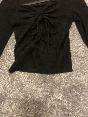 Svart långärmad topp med snörning - Svart långärmad topp med snörning och knyt framtill. Modellen är croppad och har en mjuk, stickad känsla. Perfekt för dig som gillar trendiga detaljer och vill ha en topp som sticker ut lite extra.