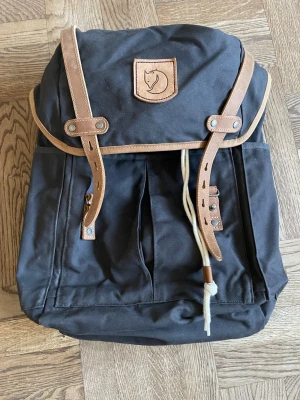 Svart Fjällräven ryggsäck G-1000 - Klassisk svart ryggsäck från Fjällräven i slitstarkt G-1000-material med detaljer i brunt läder. Ryggsäcken har lock med läderremmar, dragskostängning och justerbara axelband. Ikoniska Fjällräven-loggan framtill. Perfekt i alla lägen; till skola, jobb, träning eller utflykter. Tack vara tygsnörkkngen upptill kan volymen justeras. Knappt använd; som ny!