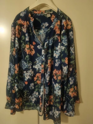 Blommig blå blus med långa ärmar - Snygg blus i mörkblått med färgglatt blommönster i orange, vitt och grönt. Luftig passform med v-ringning och långa ärmar. Perfekt för dig som gillar en bohemisk och avslappnad stil.