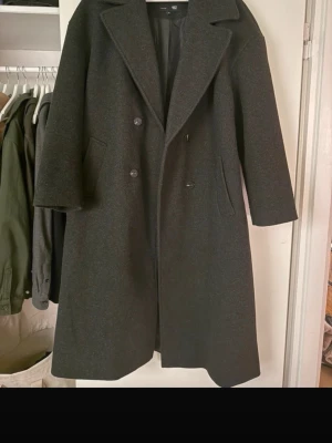 Mörkgrå kappa  - Stilren kappa . Dubbelknäppt framtill med breda slag och två snedställda fickor. Klassisk rak passform som passar perfekt till höst och vinter. Snygg och tidlös design.