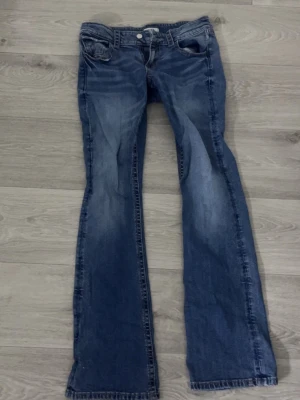 Blå bootcut jeans  - Säljer ett par blå bootcut jeans med snygga slitningar och klassisk femficksdesign. Jeansen har ljusare partier framtill och bakfickor med knappdetaljer. Perfekta för en avslappnad och trendig look.