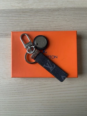 Vuitton nyckelring i svart läder - Snygg nyckelring från  Vuitton i svart präglat läder med silverfärgade metalldetaljer. Har både rund ring och karbinhake samt en rund bricka med LV-logga. Perfekt accessoar för att hålla koll på dina nycklar och ge en lyxig touch till din vardag.