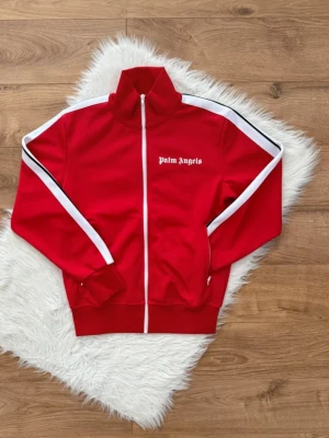 Röd Palm Angels zip hoodie - Snygg röd hoodie från Palm Angels med vit dragkedja och vita ränder längs ärmarna. Loggan är tryckt i vitt på bröstet. Hoodien har hög krage och ribbade muddar. Perfekt för en streetwear-look.