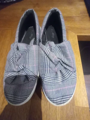 Skor med knutdetalj - Säljer ett par loafers från Alleh i grått rutigt tyg med svarta och vita detaljer samt diskreta rosa linjer. Skorna har en snygg knutdetalj framtill och vit platåsula som ger extra höjd. Perfekta för dig som gillar trendiga och bekväma skor med unik design.