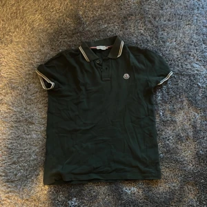 Moncler piké - Snygg mörkgrön pikétröja från Moncler med klassisk krage och vita ränder på krage och ärmslut. Tröjan har korta ärmar, knappar framtill och Moncler-logga på bröstet. Perfekt för en stilren och sportig look.