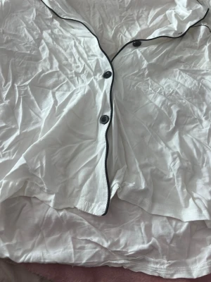 Vit pyjamaströja med svarta knappar - Klassisk vit pyjamaströja med svarta knappar och svart passpoal längs kanterna. Skön och luftig modell med kort ärm, perfekt för avslappnade kvällar hemma. Tillverkad i mjukt tyg som känns behagligt mot huden. Tyvärr lite skrynklig med det går att stryka, storlek L men skulle säga att den sitter mer som S- M 