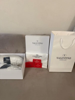Valentino Garavani vita sneakers - Snygga vita sneakers från Valentino Garavani med svart detalj över snörningen. Tillverkade i slätt läder med guldfärgad logga på plösen. Klassisk låg modell med stilren design och vita skosnören. Perfekta för dig som gillar exklusiva och trendiga skor.