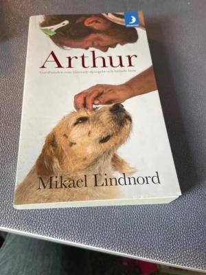 Arthur av Mikael Lindnord - En gripande pocketbok med titeln 'Arthur' av Mikael Lindnord. Omslaget visar en man som klappar en hund, vilket speglar bokens tema om vänskap mellan människa och hund. Boken har vit framsida med färgbild och röd text.