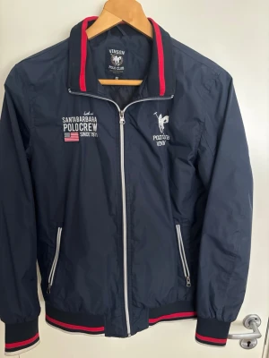 Marinblå vindjacka från Vinson Polo Club - Snygg marinblå vindjacka från Vinson Polo Club med vita och röda detaljer. Jackan har dragkedja, två fickor med dragkedja och broderade loggor samt text på bröstet. Mycket bra skit, använd 5-6 gånger 