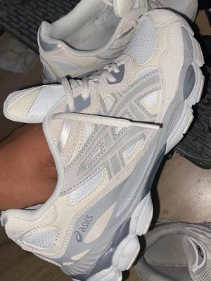 Vita och grå Asics sneakers - Snygga Asics sneakers i vitt och ljusgrått med mesh och mockadetaljer. Klassisk låg modell med tydliga Asics-loggor på sidan och bekväm, vadderad plös. Perfekta för dig som gillar stilrena och sportiga skor.