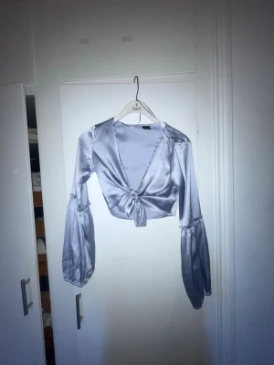 Ljusblå omlottopp i satin - Trendig silvrig omlottopp i satin med långa puffiga ärmar och knyt framtill. Croppad modell med v-ringad urringning som ger en snygg och modern look. Croptop