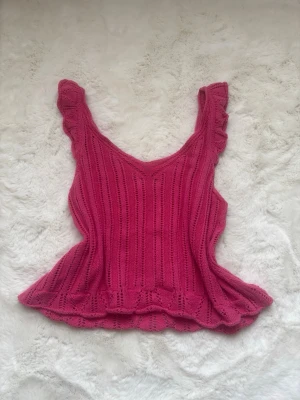 Stickad rosa linnetopp med volang - Supersöt stickad rosa linnetopp med volangkant och breda axelband. Toppen har ett ribbat mönster och är perfekt för dig som gillar en feminin och trendig stil. Snygg att matcha med jeans eller kjol.