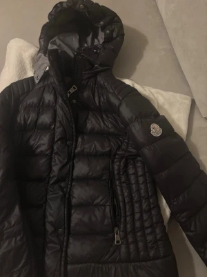 Svart dunjacka från Moncler med huva - Snygg svart dunjacka från Moncler med quiltad design och avtagbar huva. Jackan har dragkedja och knappar framtill samt två fickor med dragkedja. Moncler-logga på ärmen ger en exklusiv touch. Perfekt för kalla vinterdagar.