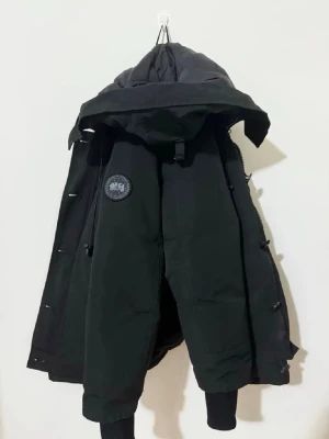 Svart jacka från Canada Goose - Snygg svart dunjacka från Canada Goose med huva och klassisk patch på ärmen. Jackan har dragkedja, knappar och ribbade muddar vid ärmslut. Perfekt för kalla vinterdagar och har en stilren, tidlös design.