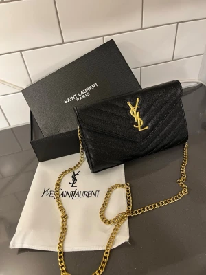 Svart YSL väska - Snygg svart kuvertväska från Saint Laurent med ikoniskt YSL-emblem i guld framtill. Väskan har en elegant kedjerem i guld och är tillverkad i läder med diskret mönster. Perfekt för dig som vill ha en lyxig accessoar med stilren design.