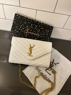 Vit YSL väska - Snygg vit kuvertväska från Yves Saint Laurent med klassiskt YSL-emblem i guld på framsidan. Väskan har quiltat mönster och en elegant guldfärgad kedjerem. Perfekt för att lyfta vilken outfit som helst.