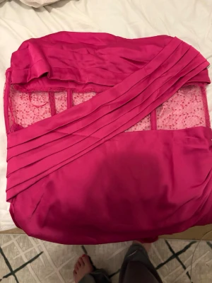 Rosa långklänning med spetsdetaljer - Snygg långklänning i stark rosa färg med transparent spets upptill. Klänningen har omlottdetaljer framtill och en slits där fram.  Materialet är glansigt och känns lyxigt, perfekt för speciella tillfällen. Oanvänd klänning från hello molly. Storlek Xl men har man större byst passar de bättre till nån som har L. 