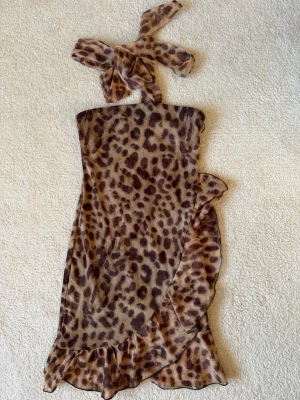 Leopardmönstrad kort klänning med volang - Säljer en leopardmönstrad kort klänning med volangkant och knyt i halsen. Klänningen har en figurnära passform och är axelbandslös med ett dekorativt omlottband. Perfekt för dig som vill sticka ut med ett djurmönster!