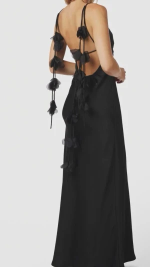 Colly Heavy Sateen Dress Black - Elegant svart långklänning med tunna axelband och öppen rygg. Klänningen har dekorativa blommor längs banden och sidorna samt v-ringad front. Tillverkad i ett glansigt sateen material som ger en lyxig känsla. Perfekt för dig som vill sticka ut. Använt 1 gång 