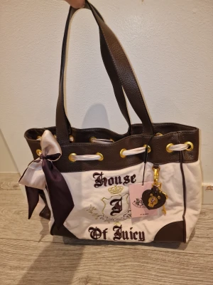House of Juicy Couture daydreamer tote  - Snygg handväska från Juicy Couture i brunt och vitt tyg med detaljer i konstläder. Väskan har broderad text, guldfärgade öljetter, rosett och hjärtformad detalj. Perfekt för dig som gillar statement-accessoarer och unika detaljer.