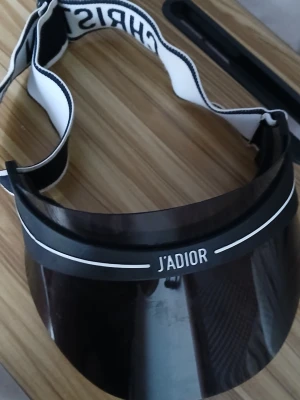 J'ADIOR solskärm från Dior - Cool svart solskärm från Dior med texten J'ADIOR framtill och elastiskt band med Christian Dior-logga. Skärmen är tillverkad i plast och har vita detaljer. Perfekt accessoar för att skydda ansiktet mot solen med stil.