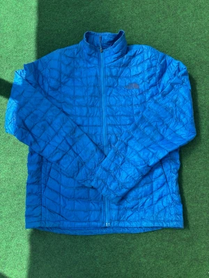 Blå pufferjacka från The North Face - Snygg blå pufferjacka från The North Face med quiltat rutmönster och hög krage. Jackan har små slitage som bilderna visar! Men plagget funkar helt perfekt fortfarande 
