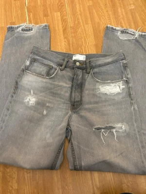Grå jeans - asos - Säljer ett par grå jeans med snygga slitningar och hål på framsidan. Jeansen har raka ben och råa kanter vid bensluten för en trendig look. Klassisk femficksmodell och knappgylf. Perfekta för dig som gillar en avslappnad och edgy stil. Kolla bild för exakt storlek