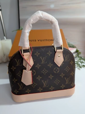Iouis Vuitton Alma BB  - En elegant handväska i klassiskt Monogram-canvas med detaljer i ljust vachetta-läder. Väskan har den ikoniska strukturerade kupolformen och är utrustad med dubbla handtag samt en matchande clochette (nyckelhållare).