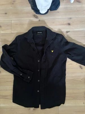 Skjorta från Lyle & Scott - Svart skjorta från Lyle & Scott med klassisk krage och knappar framtill. Skjortan har ett broderat gult örnmärke på bröstet och är långärmad. Materialet är bomull och passformen är normal.