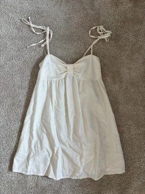 Vit kort klänning med rosett - Söt vit kortklänning med tunna axelband som knyts och en dekorativ rosett framtill. Klänningen har en luftig passform och är perfekt för varma dagar💓